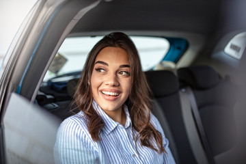 Fototapeta na wymiar Businesswoman in car on the way to her workplace. Beautiful girl in the backseat. Beautiful businesswoman is sitting on back seat of the car. She is looking through the window dreamingly and smiling.