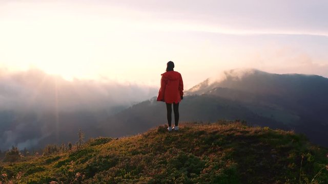 Inspired young woman traveler standing on hilltop watching spectacular mountain sunset scenery. Beautiful mountain range covered with green pine woods. Nature and freedom concept.