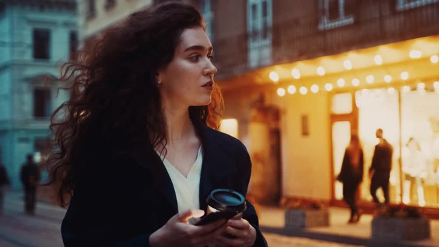 Stylish young european curly haired woman walking along the street using phone, texting in the city at night. Warm spring or autumn evening business outdoor, lifestyle girl town Slow motion close up