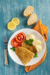 Vertical top view of a plate of healthy baked battered fish, seasoned with fresh parsley and lemon and accompanied by cooked potatoes, tomato and freshly baked bread. Blue background.