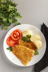 Top view of a healthy baked battered fish seasoned with fresh parsley and lemon and accompanied by cooked potatoes and tomato. Concept of nutritious food for a healthy diet. Wellness and health care.