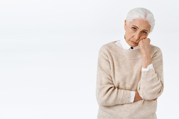 Bored or annoyed senior lady with grey combed hair, lean face on fist, look from under forehead distressed with frustration and bad mood, sulking over white background, insulted or disappointed