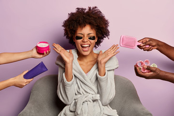 Facial skin care concept. Pretty overjoyed curly woman wears face patches, reduces wrinkles under eyes, enjoys various cosmetic treatments at same time after taking bath, being at home or spa salon