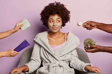 Health and beauty concept. Thoughtful young lady does spa procedures for skin health, applies facial clay mask, looks happily aside on someones hands with cosmetic products being treated by beautician