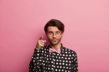 Fototapeta premium Image of handsome unshaven man points at camera, looks directly at you, wears round big spectacles, shirt and bowtie, chooses something, isolated on pink background. Facial expressions concept