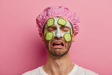 Beauty procedure, cosmetology and skin care concept. Melancholy dissatisfied man cries from negative emotions, wears bath cap, cucumbers on face, reduces fine lines, isolated over rosy wall.