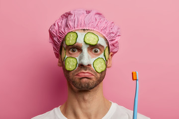 Headshot of shocked man cleanses facial skin, holds toothbrush, shower cap, ready for cleaning teeth, has serious strict look, models against pink background, visits beauty salon, has spa treatments