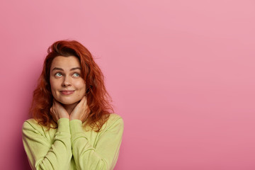 Fototapeta premium Dreamy woman in love recalls perfect date with boyfriend, thinks about cute sweet things, keeps hands on neck, looks somewhere, wears green sweater, isolated over pink wall, copy space area.