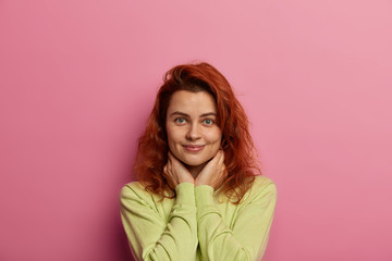 Fototapeta premium Good looking calm woman with natural beauty, ginger hair, looks with satisfaction, keeps hands under face, dressed in casual jumper, pleased to hear something good, isolated on pink background
