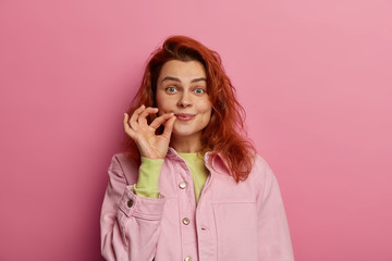 Indoor shot of secret redhead young woman zips her mouth shut, promises to keep secret, has gossip safe, holds fingers near cheek, makes promise, hides information. Secret locked in my mouth