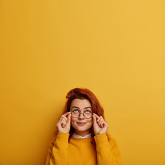 Indoor image of thoughtful redhead Caucasian woman keeps hands on frame of glasses, tries to see something above, has curious glad expression, wears yellow jumper. People, daydreaming concept