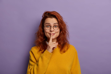 Pleased woman shushing with index finger over lips, tells confidential inforamtion, secret and gossips, wears round glasses, yellow jumper, isolated over purple background. Do not make noise