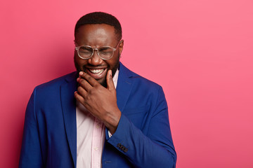 Photo of overjoyed Afro American man laughs at funny story, cant stop giggling, has white teeth, thick beard, wears formal suit, squints face, isolated on pink background, glad to achieve success - obrazy, fototapety, plakaty