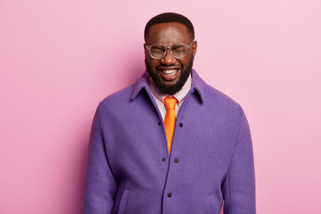Portrait of dark skinned male employee has hysterical laugh, positive mood, smiles broadly, wears purple jacket, orange tie, white shirt hears hilarious joke, isolated over pink pastel background