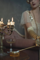 Retro 1920s fashion woman ticking cigarette in ashtray while sitting on sofa beside table with candlestick.