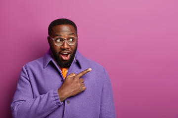 Expressive surprised black man points right with index finger and says wow, sees shocking things, wears purple jacket, isolated over lilac background, copy space area. Advertisement and promo