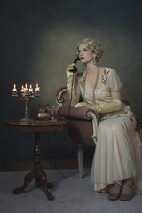 Retro 1920s fashion woman on the phone while sitting on sofa beside table with candlestick and...
