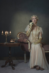 Retro 1920s fashion woman on the phone while sitting on sofa beside table with candlestick and glass of champagne.
