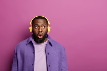 Listening concept. Surprised black man has thick bristle, looks shocked away, uses best music app...