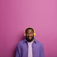 Obraz premium Studio shot of happy unshaven man listens audio track in headphones, looks above with smile, dressed in purple shirt, has healthy dark skin, isolated over lilac background, gets music subscription