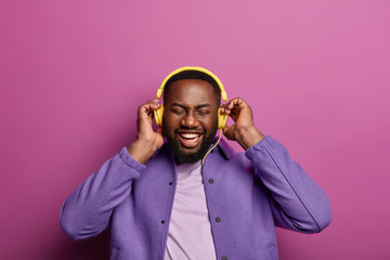 Obraz premium Music fan concept. Smiling black man with dark bristle, uses musical accessory gadget, enjoys perfect sound in headphones, has broad smile, white teeth, closes eyes, dressed in fashionable clothes