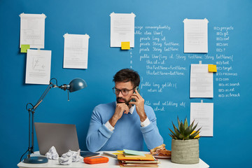 Busy male freelancer in blue outfit, checks information on laptop computer, uses modern cellphone gadget for calling, sits at white table with papers, reads news or watches video, download new app