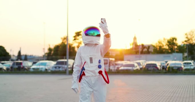 Astronaut in the full equipped costume and head armour standing at the cars parking while taking selfie photos on the smartphone or having videochat via web cam. Outdoor.
