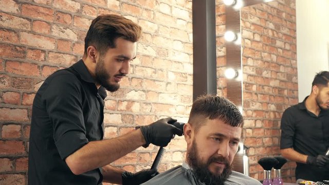Young barber trimming hair to handsome bearded client in barbershop. Hairdresser holding electric clipper and cutting hair of male client sitting in chair at barber shop