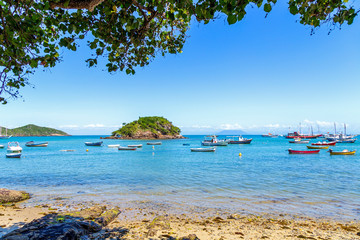 Seascape of Armacao Beach in Buzios, Rio de Janeiro, Brasil. Panoramic view of the bay. Boats and sailboats anchored in the sea. Tree leafs, clear water, blue sky, some rocks and a island.