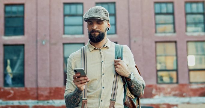 Smiled Caucasian young hipster guy with a beard and in a cap standing outdoors and texting a message on the smartphone.