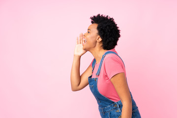 African american woman with overalls over isolated pink background shouting with mouth wide open