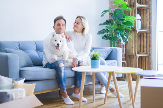 Young beautiful couple with dog sitting on the sofa at new home around cardboard boxes