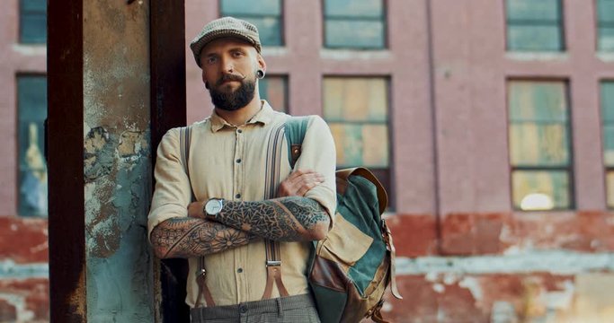 Portrait shot of the handsome stylish Caucasian hipster guy with tattoos and beard posing to the camera and crossing hands in front of him. Outside.
