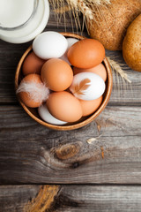 Fresh white and brown chicken eggs in a wooden bowl, milk, bread, and wheat ears. Raw eggs on wooden background