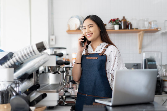 Happy Asian Woman Barista Working On Laptop Smiling And Talking On Smartphone, Accepting Writing Order At A Coffee Shop. Waitress In Cafeteria Small Business Owner, Online Purchase And Service Concept