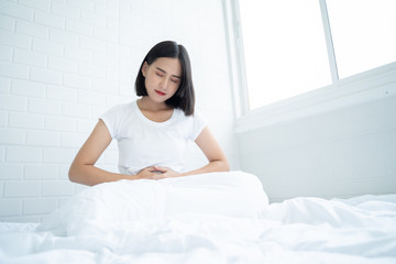 Asian woman unhappy lying on the bed looking sick, suffers from stomach ache in the bedroom, stomachache because of menstruation and eating spoiled food, Chronic gastritis. Abdomen bloating concept