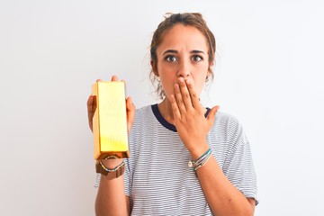 Young beautiful redhead woman holding gold ingot over isolated background cover mouth with hand shocked with shame for mistake, expression of fear, scared in silence, secret concept