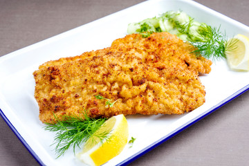 Traditionelles frittiertes Wiener Schitzel von der Oberschale vom Kalb mit Gurken Salat und Zitronenscheiben angeboten als closeup auf einem weißen Tablett