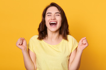 Close up portraiot of happy young woman with beautiful dark hair, clenching fist while looking directly at camera, wearing casual t shirt, isolated over yellow background. People emotions concept. - obrazy, fototapety, plakaty