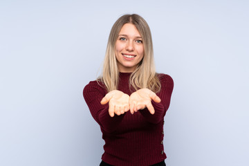 Young blonde woman over isolated blue background holding copyspace imaginary on the palm to insert...