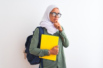 Young Arab student woman wearing hijab and backpack holding a book over isolated background serious face thinking about question, very confused idea