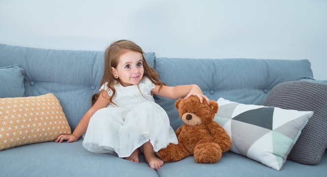 Adorable blonde toddler sitting on the sofa playing with teddy bear at home