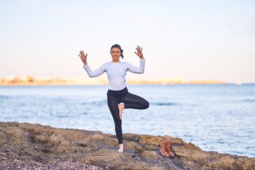Young beautiful sportwoman smiling happy practicing yoga. Coach with smile on face teaching tree pose at the beach