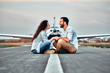 Beautiful romantic couple is sitting on runway private plane in airport. Handsome man and...