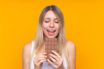 Young blonde woman over isolated blue background eating a chocolate tablet
