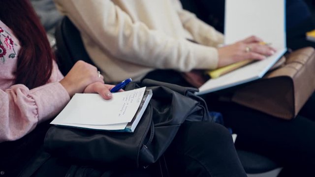 Women sitting in a row during the conference. Young females hold copybooks and write important information during the seminar. Close-up.