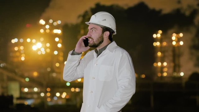 Young angry engineer in helmet and white robe shouting on phone standing by gas refinery.