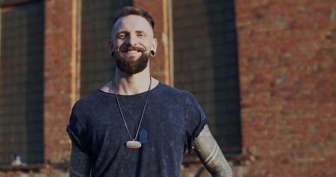 Portrait shot of the attractive and stylish young Caucasian male hipster with beard turning face to the camera and looking straight. Outside at the brick wall background.