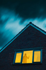 Burglar with pistol in eerie house with illuminated window under stormy sky at dusk.