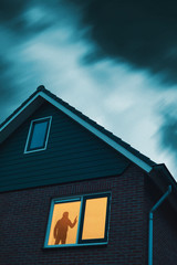 Intruder with handgun in eerie house with illuminated window under stormy sky at dusk.